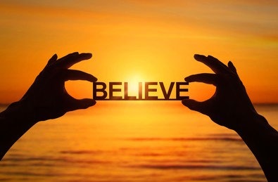 Silhouetted hands holding the word BELIEVE against a sunset